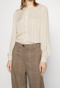 Donna che indossa una camicia crema con bottoni e dettagli plissettati, maniche lunghe, abbinata a pantaloni a pied de poule marroni a vita alta.