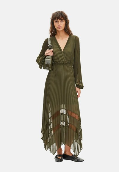 Woman wearing a long olive green pleated dress with sheer patterned panels, black studded loafers, and carrying a matching shoulder bag.