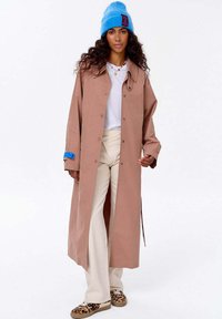 Kobieta w długim brązowym płaszczu, białej bluzce, kremowych spodniach, butach w panterkę i jasnoniebieskiej czapce z czerwoną łatą, stojąca na białym tle.