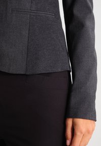 Ciemnoszary żakiet o dopasowanym kroju z subtelną teksturą, jedną kieszenią, slim fit oraz gładkim wykończeniem, zestawiony z czarnymi spodniami.