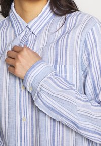 Camicia a righe blu e bianche con colletto, dotata di tasca e tessuto testurizzato. Mostra il polsino e una mano che sistema il colletto.
