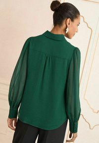 Blusa verde con maniche a sbuffo trasparenti e colletto. Presenta bottoni sulle maniche e una vestibilità morbida.