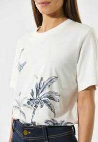 Hvid bomulds t-shirt med blå botaniske og sommerfugletryk. Kortærmede og med rund halsudskæring, hjulpet af mørke denimbukser.
