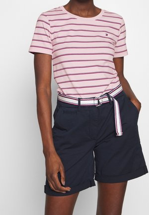 Kvinde iført en rosa striped T-shirt og marineblå shorts med opbukket kant og et stofbælte i hvidt, marineblåt og rødt.