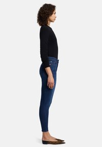 Jean skinny en denim bleu foncé avec une texture lisse, associé à un pull noir ajusté et des ballerines imprimées léopard. Vue de côté.