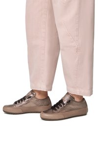 Metallisch bronze Sneakers mit glatter Textur, runder Zehe, kontrastierenden schwarzen Schnürsenkeln und einer gepolsterten Sohle, kombiniert mit hellrosa Hosen.