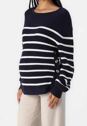 Personne enceinte portant un pull en maille rayé bleu marine et blanc avec des boutons latéraux et un pantalon côtelé beige clair.