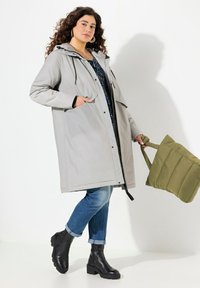 Parka impermeabile grigio chiaro con cappuccio, tasche frontali e chiusura a pressione, indossata sopra un top con fantasia e jeans blu, abbinata a stivali neri e una borsa a tracolla verde.