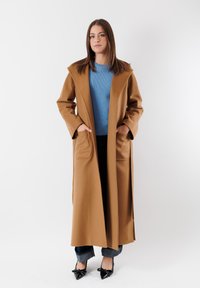 Lungo cappotto di lana marrone con ampio colletto e tasche anteriori, indossato su un maglione blu a coste e jeans scuri, abbinato a scarpe con tacco nere.