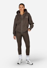 Barna cipzáras kapucnis pulóver elülső zsebbel, hozzá illő barna leggings-szel. Fehér logós díszek a ruhán és a zoknikon, valamint fehér sportcipő.