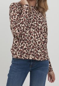 Top à manches longues en imprimé léopard crème, avec des taches noires et bordeaux. Tissu doux avec une coupe décontractée et un col arrondi. Jean en denim visible.