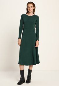 Frau trägt ein langärmliges, dunkelgrünes Midi-Kleid mit taillierter Passform, kombiniert mit schwarzen, knöchellangen Lederstiefeln vor einem schlichten Hintergrund.