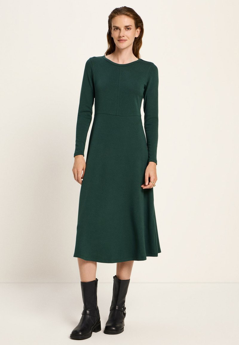 Frau trägt ein langärmliges, dunkelgrünes Midi-Kleid mit taillierter Passform, kombiniert mit schwarzen, knöchellangen Lederstiefeln vor einem schlichten Hintergrund.