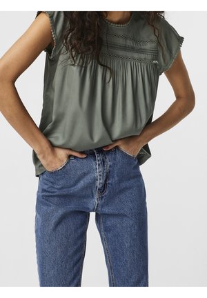 Kvinde iført en ærmeløs olivengrøn bluse med blondedetaljer og højtaljede blå jeans, med hænderne puttet i forlommerne.