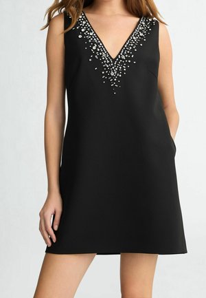 Vestido negro sin mangas con escote en V profundo adornado con abalorios y lentejuelas plateadas, llevado por una mujer con la mano en el bolsillo.