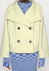 Dvojřadý kabát ze světle žluté vlněné směsi, s širokým límcem, černými knoflíky a bočními kapsami. Nalezeno přes modrý vzorovaný pletený top.