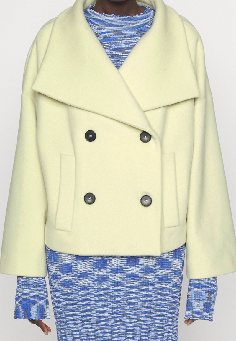 Dvojřadý kabát ze světle žluté vlněné směsi, s širokým límcem, černými knoflíky a bočními kapsami. Nalezeno přes modrý vzorovaný pletený top.