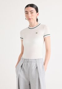 Weißer Kurzarm-Body mit grünen Akzenten und kleinem Logo, kombiniert mit hellgrauen Jogginghosen mit Kordelzug, die Falten und Seitentaschen aufweisen.