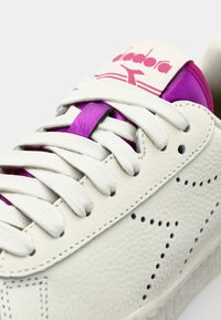 Hvit teksturert lær sneakers med lilla detaljer, flate hvite lisser og en rosa logo på tungen med perforerte detaljer på siden.