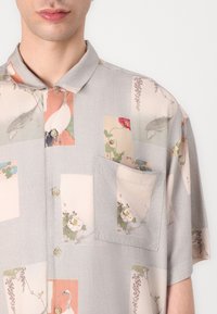 Cămașă cu mânecă scurtă, de culoare gri deschis, cu un model floral și animal print. Dispune de un buzunar pe piept și de un guler clasic.