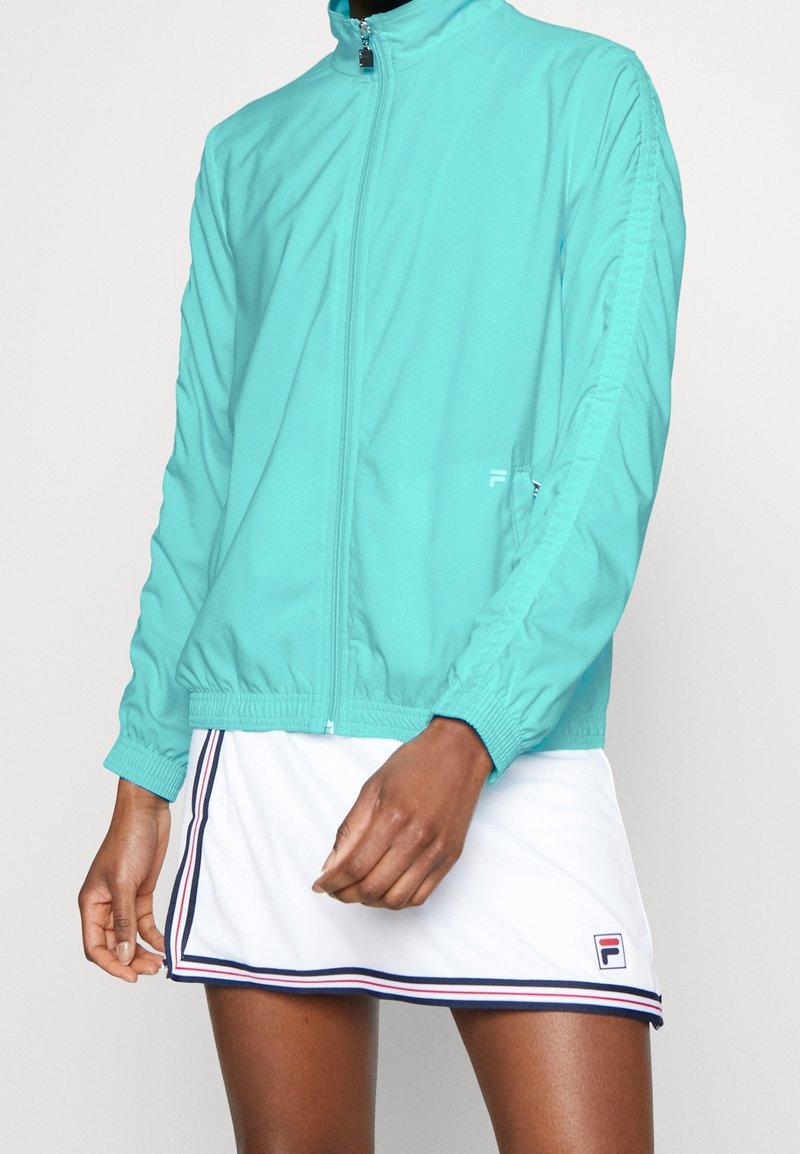Veste zippée turquoise légère avec poignets et ourlet élastiques, arborant un logo discret. Associée à une jupe blanche ornée de rayures bleu marine et rouges.