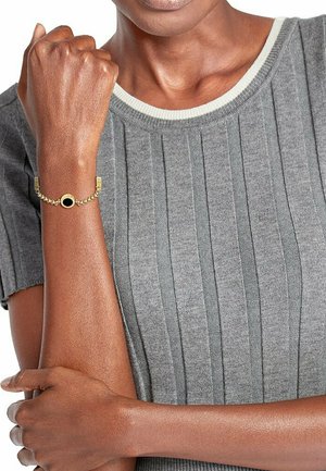 Gouden kettingarmband met een ronde zwarte accent, gedragen om een pols met een gestructureerde grijze geribbelde top.