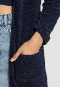 Mörkblå stickad cardigan med långa ärmar, med en ficka fram. Tyget har en mjuk, strukturerad yta.