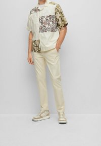Camicia a maniche corte con motivi misti in beige, marrone e grigio, abbinata a pantaloni beige chiaro e scarpe sportive in tonalità tenui.