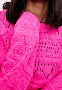 Jasnoróżowy, dziergany sweter z dużymi teksturowanymi wzorami i otwartymi oczkami. Ściągacze na mankietach i dekolcie dodają szczegółów do projektu.