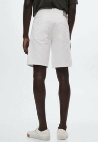 Hombre vestido con pantalones cortos blancos hasta la rodilla, camiseta oscura y zapatos blancos tipo slip-on, de pie contra un fondo claro y uniforme.
