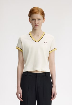 Jeune personne aux cheveux roux courts portant un t-shirt blanc à col en V avec un bord jaune et un pantalon noir, sur un fond clair uni.