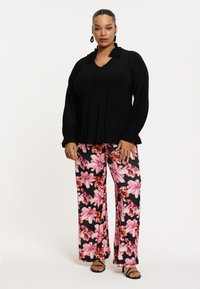 Sort langærmet top med flæsekant og v-hals, parret med blomstrede brede bukser i pink og sort. Model iført sorte sandaler.