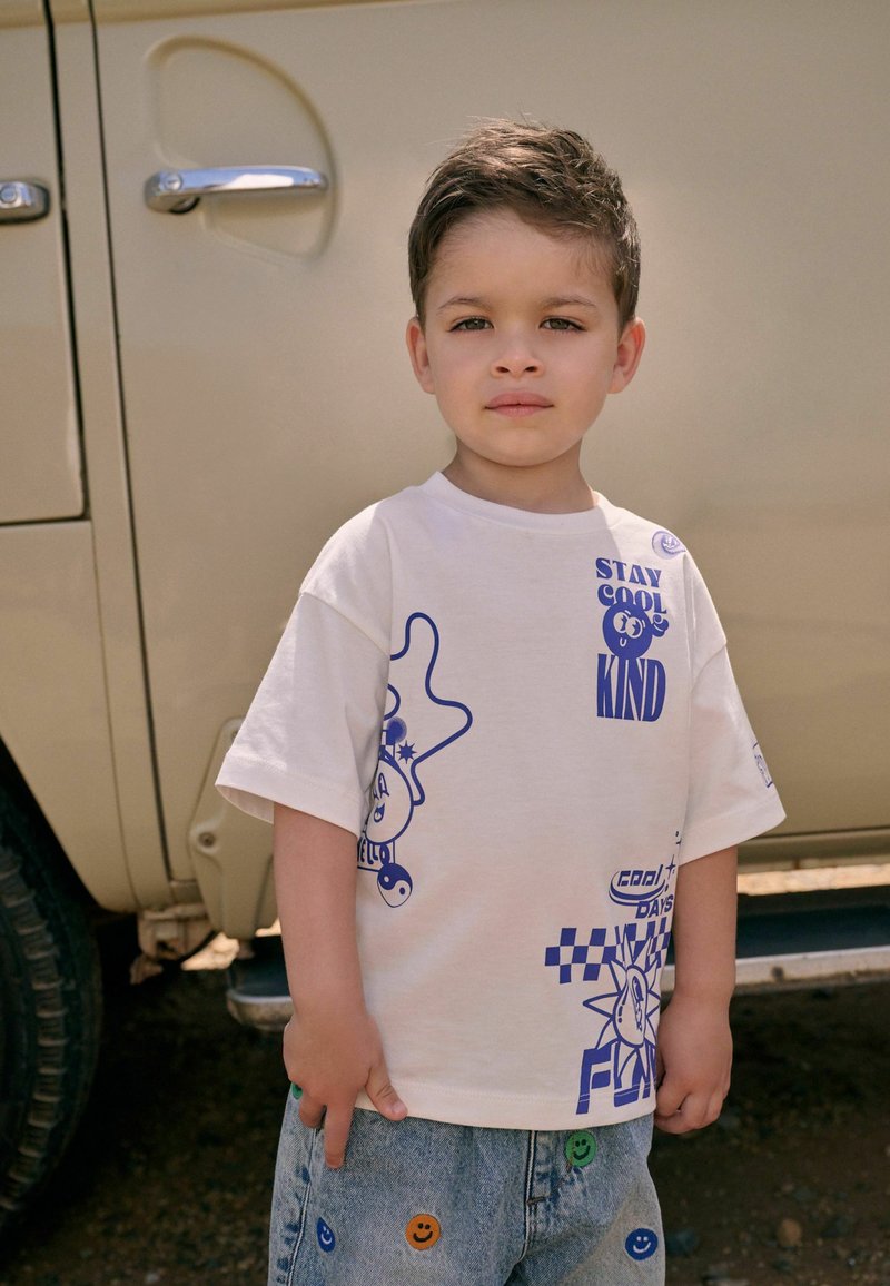 Witte katoenen t-shirt met blauwe grafische prints, inclusief tekst en speelse ontwerpen. Gepaard met lichtblauwe denim shorts met kleurrijke smiley accenten.