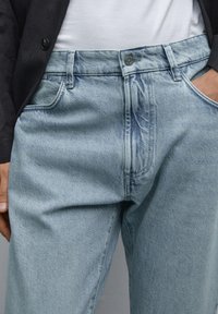 Light blue denim jeans with faded wash, classic five-pocket design, and a button closure at the waist. Smooth texture and straight cut.