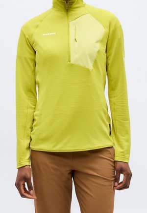 Person, die ein langärmliges, limegrünes Quarter-Zip-Top mit Brusttasche und braunen Hosen trägt, steht vor einem schlichten Hintergrund.