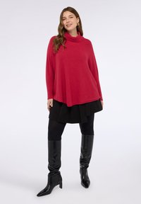 Poncho a dolcevita rosso in materiale morbido, con dettagli a coste, abbinato a una gonna nera plissettata e stivali neri con tacco alto.