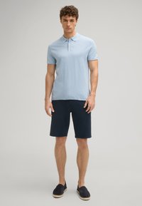 Polo shirt blu chiaro con colletto, abbinato a pantaloni corti texture navy scuro e scarpe slip-on nere. Sfondo semplice.
