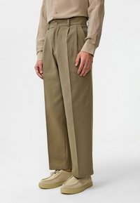 Beige geplooide broek met een hoge taille en rechte pijpen, gemaakt van gestructureerde stof, gecombineerd met lichte schoenen.