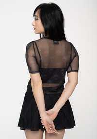 Black sheer mesh top with short sleeves, featuring a skull pattern, paired with a black pleated skirt. Model stands facing away.
