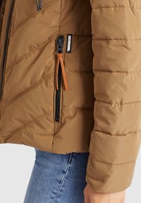 Manche de veste matelassée marron et fermeture éclair latérale avec tirette en cuir portée sur un jean en denim bleu.