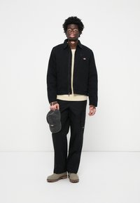Chaqueta negra de pana con cremallera, suéter beige, pantalones cargo negros, gorra gris en la mano y botas verde oliva. Logo destacado en la chaqueta.