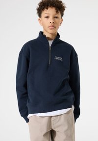 Sudadera de polar azul marino con cremallera de un cuarto, cuello redondo y detalle de texto impreso en el pecho. Textura suave y corte relajado.