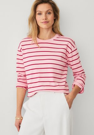 Vrouw met blond haar die een roze shirt met lange mouwen met rode horizontale strepen en een witte broek draagt, staand met één hand in de zak.