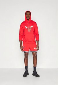 Egy férfi áll, piros Chicago Bulls kapucnis pulóvert, hozzá illő kosárlabdás rövidnadrágot, fekete Nike zoknit és fekete sportcipőt viselve, egyszínű háttér előtt.