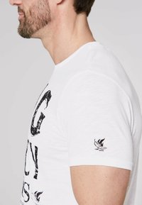 Weißes T-Shirt mit kurzen Ärmeln, das vorne einen abgenutzten schwarzen Grafiktext und ein kleines gesticktes Logo auf dem linken Ärmel zeigt.