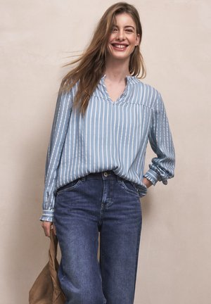 Uśmiechnięta kobieta z długimi włosami w niebiesko-białej bluzce w paski i niebieskich dżinsach, trzymająca w jednej ręce brązową torbę.