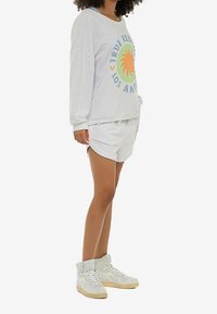 Witte oversized sweatshirt met kleurrijke zonafbeelding en tekst, gecombineerd met witte korte broeken. Draagt witte hoge sneakers.