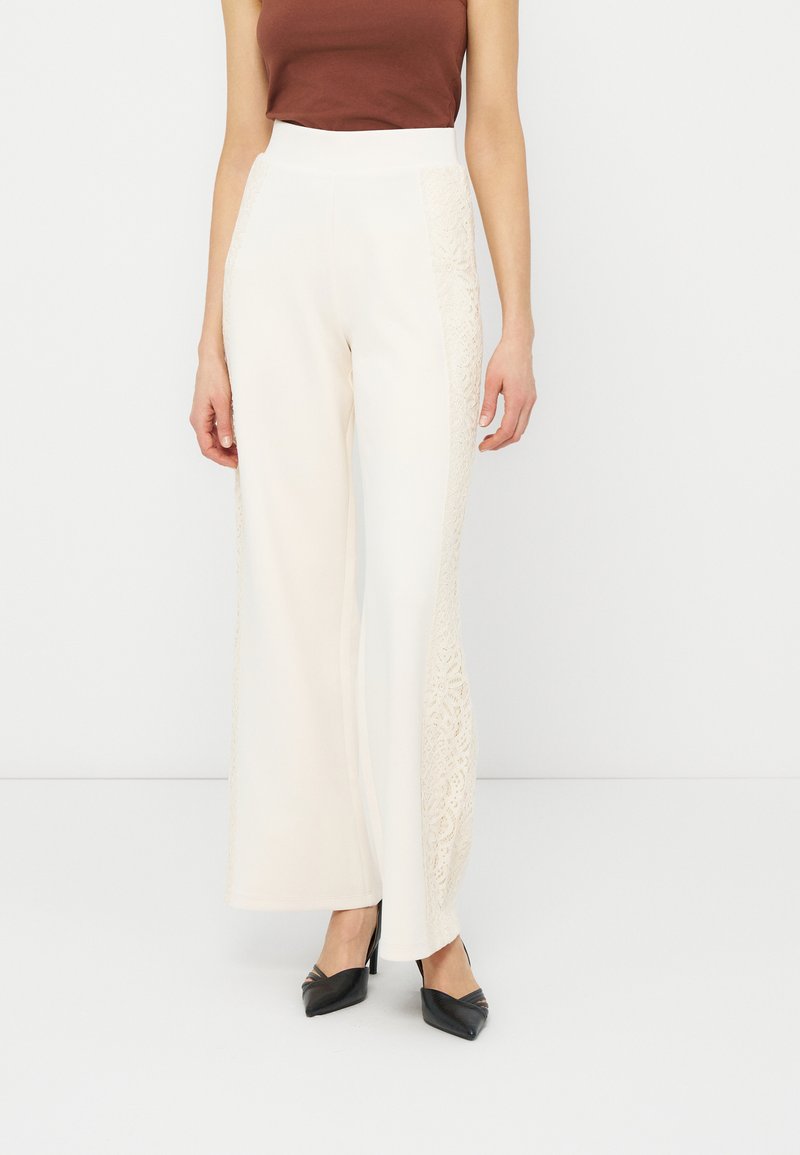 Femme portant un pantalon blanc à taille haute et jambes larges avec des panneaux latéraux en dentelle, un haut marron sans manches et des talons pointus noirs.