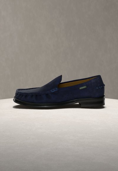 Μοκασίνι από μπλε σκούρο σουέντ με μαύρη σόλα, σχεδιασμό που φοριέται εύκολα, ραφές μοκασίνου και μικρή πράσινη ετικέτα της μάρκας στο πλάι.