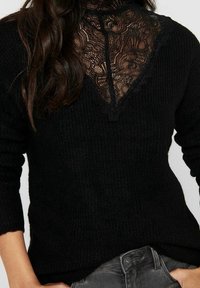 Pull noir en maille avec une superposition de dentelle au niveau de l'encolure, bordée d'un bord festonné et manches longues. Tissu texturé.